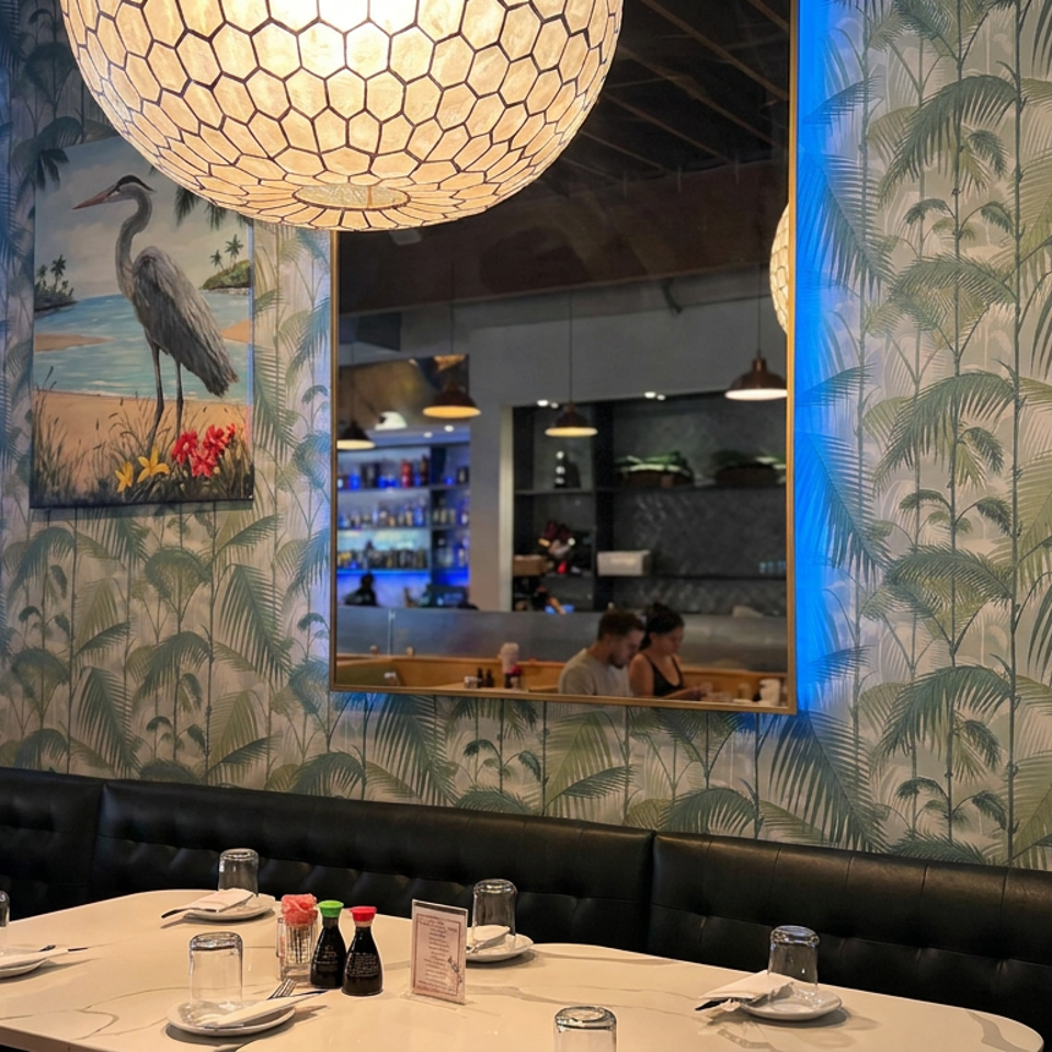 Restaurant dining area with tropical palm wallpaper, illuminated mirrors, pendant lights, black banquettes, and set tables.