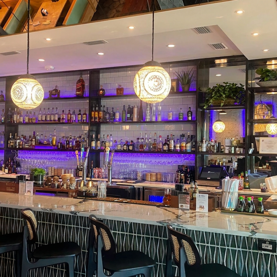 Upscale restaurant bar with purple-lit liquor shelves, marble counter, pendant lights, tropical wallpaper, and a bartender.