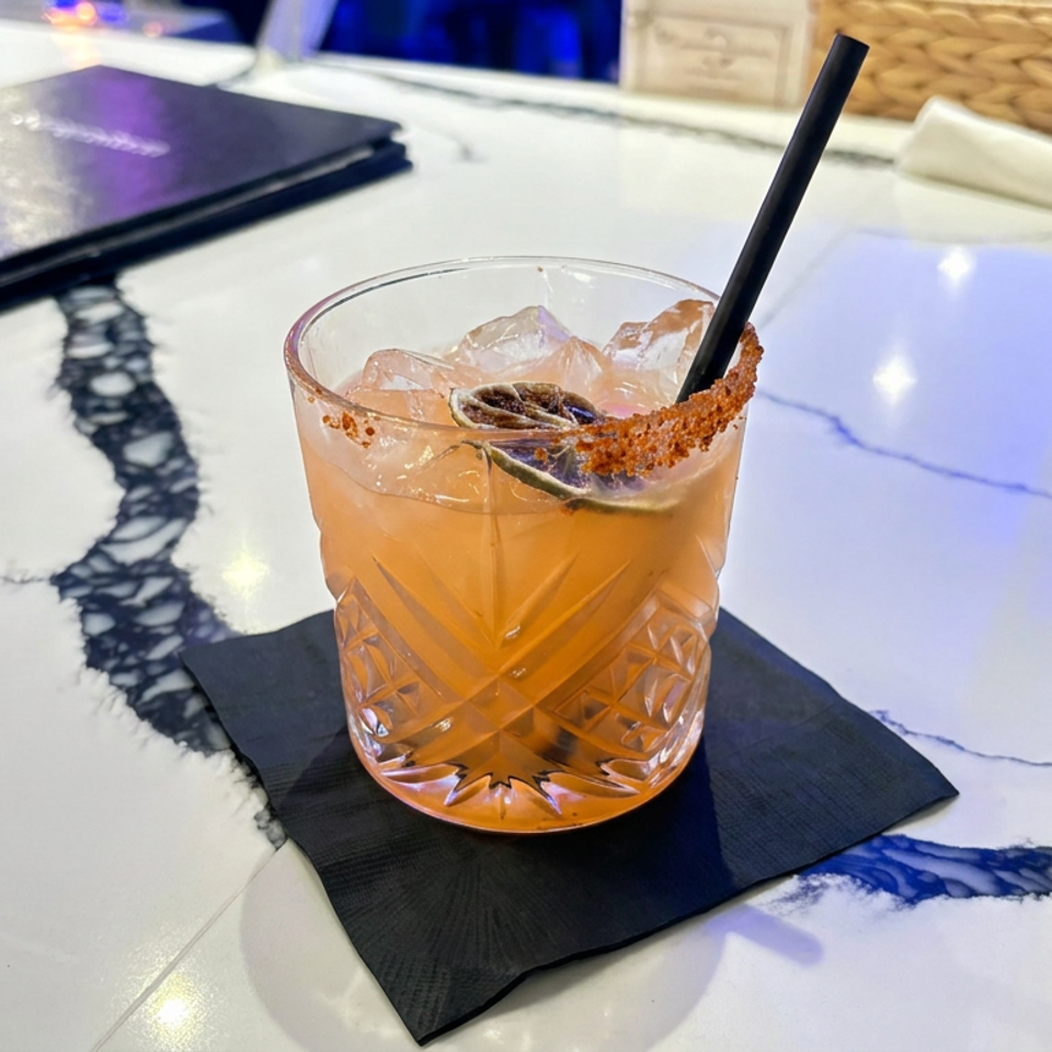 Amber cocktail with ice, black straw, and cinnamon-sugar rim served on a black napkin atop a marble restaurant table.